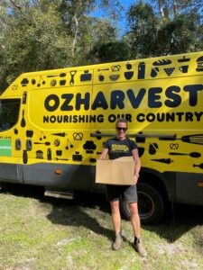 a yellow and black truck parked with OZHarvest logo on it with a woman receiving a donation standing in front of it